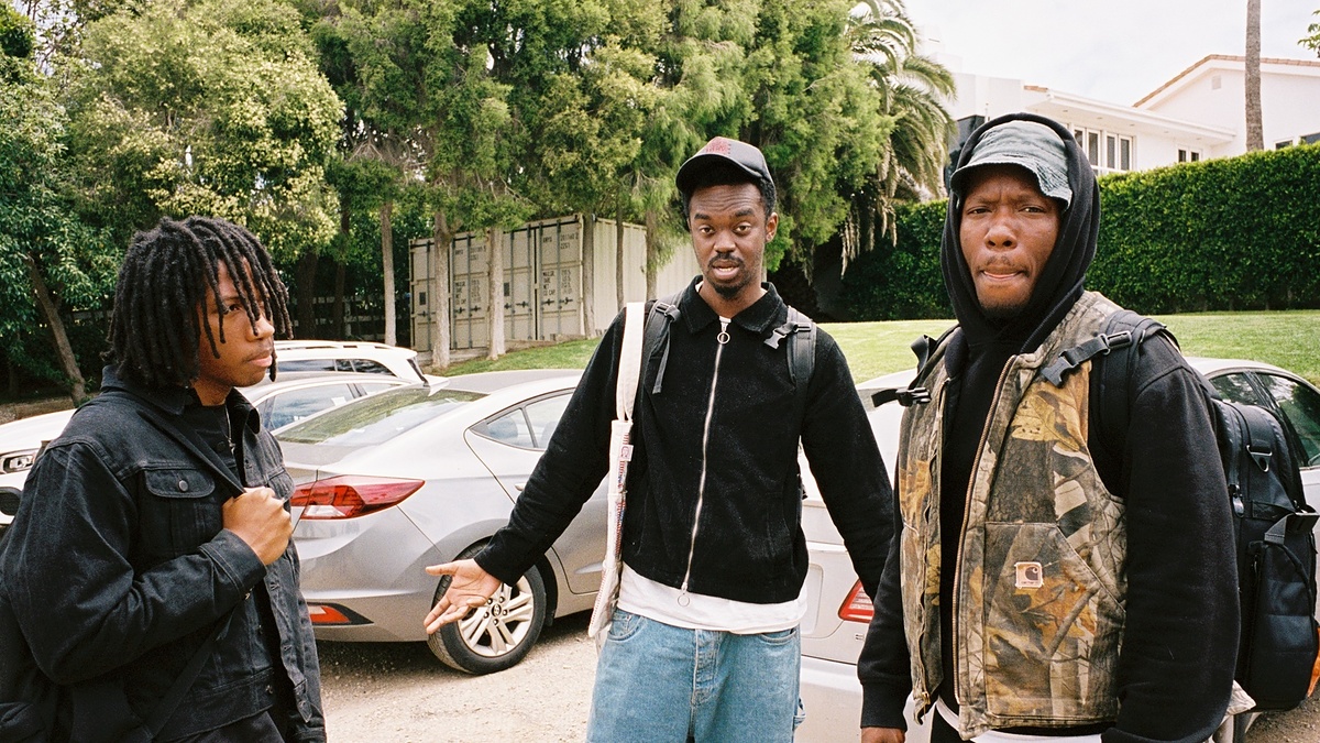 Three men stand outdoors near parked cars, dressed casually in jackets and hoodies, with trees and a house in the background.