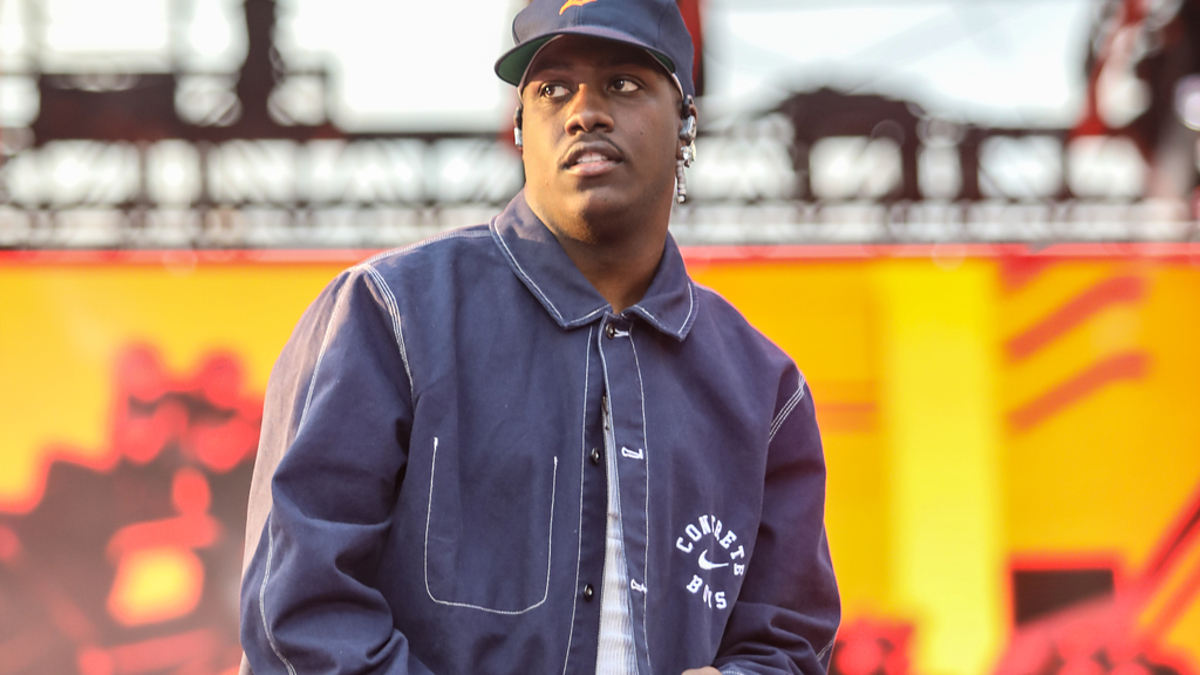 A rapper wearing a baseball cap and a blue jacket performs on stage at a music event
