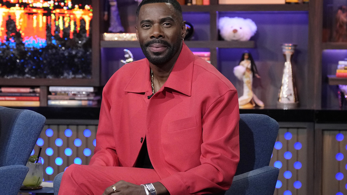 Colman Domingo sitting on a talk show set, wearing a stylish suit jacket with matching pants. Shelves with various items and a lit backdrop are visible behind him