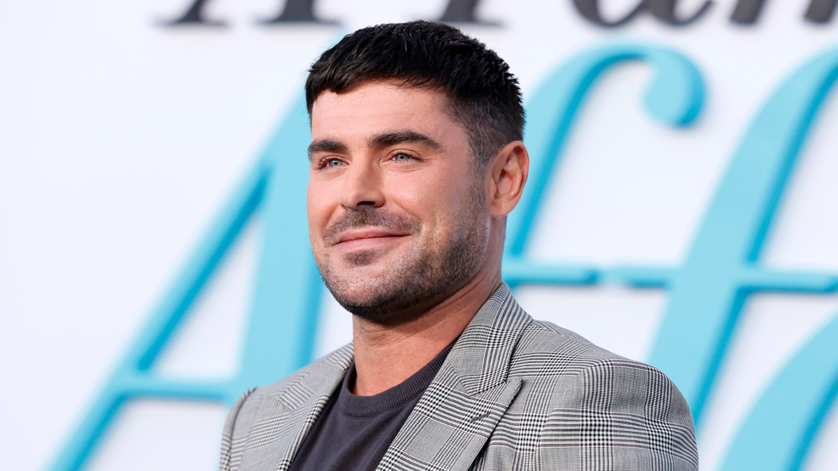 Zac Efron at a public event, wearing a checkered blazer and a black shirt, smiling in front of a backdrop with large text