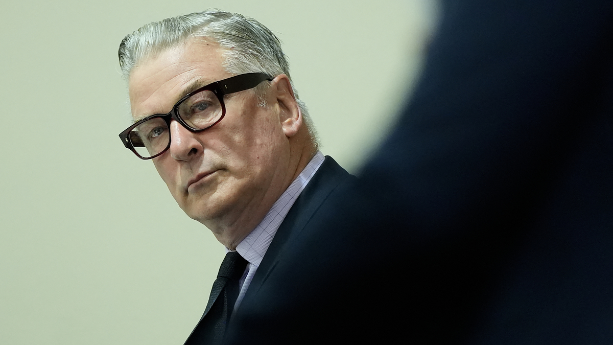 Alec Baldwin is wearing a dark suit and glasses while looking off to the side during an event