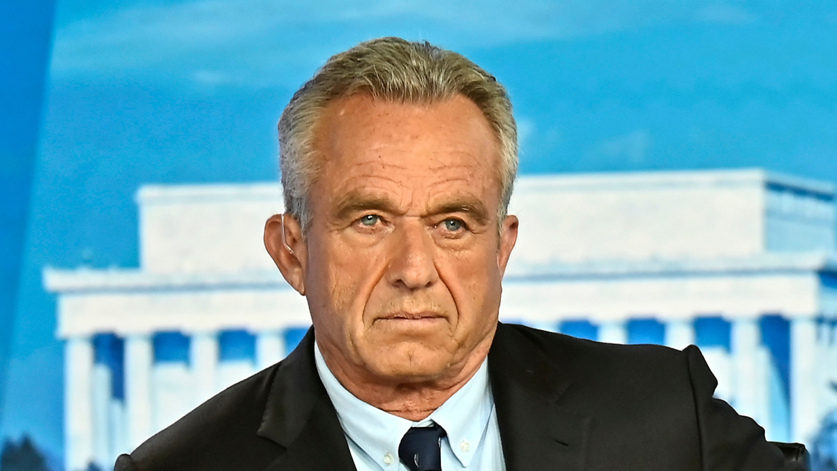 A man wearing a dark suit and light-colored shirt with a tie is in front of a building with columns