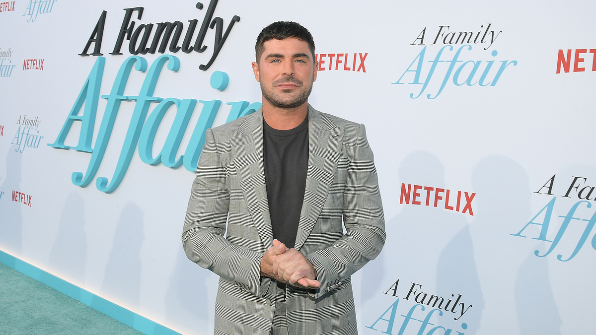 Zac Efron, in a checked suit, stands in front of a backdrop that reads "A Family Affair" with Netflix branding