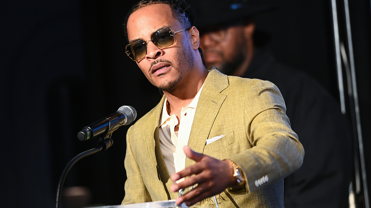 Man in a tan suit and sunglasses speaks at a podium with a microphone