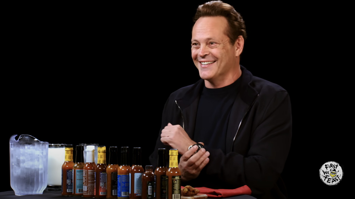 Vince Vaughn smiling while seated at a table with various hot sauces, a pitcher of water, and a milkshake on the set of "Hot Ones"