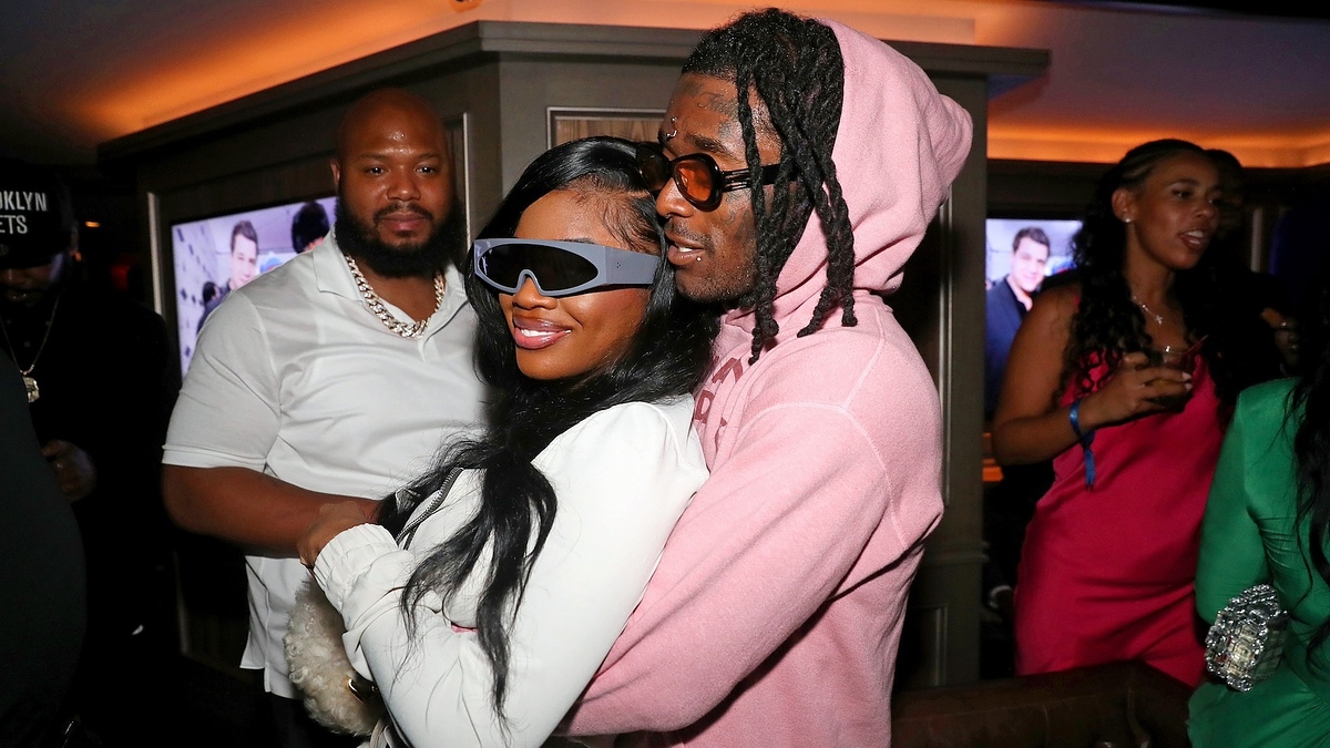 JT in futuristic sunglasses and Lil Uzi Vert in a pink hoodie, pose together at an event while a security guard and other guests look on