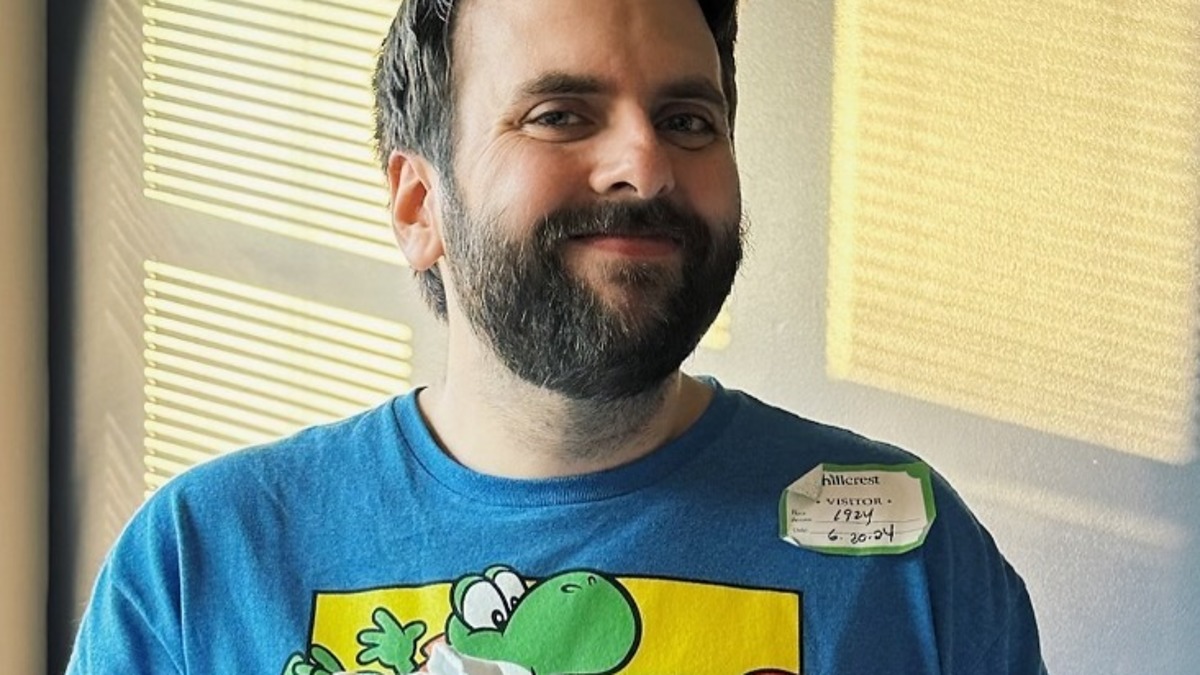 Man wearing a Super Mario Yoshi shirt holds a newborn baby wrapped in a hospital blanket, with a visitor sticker reading "8-20-24"