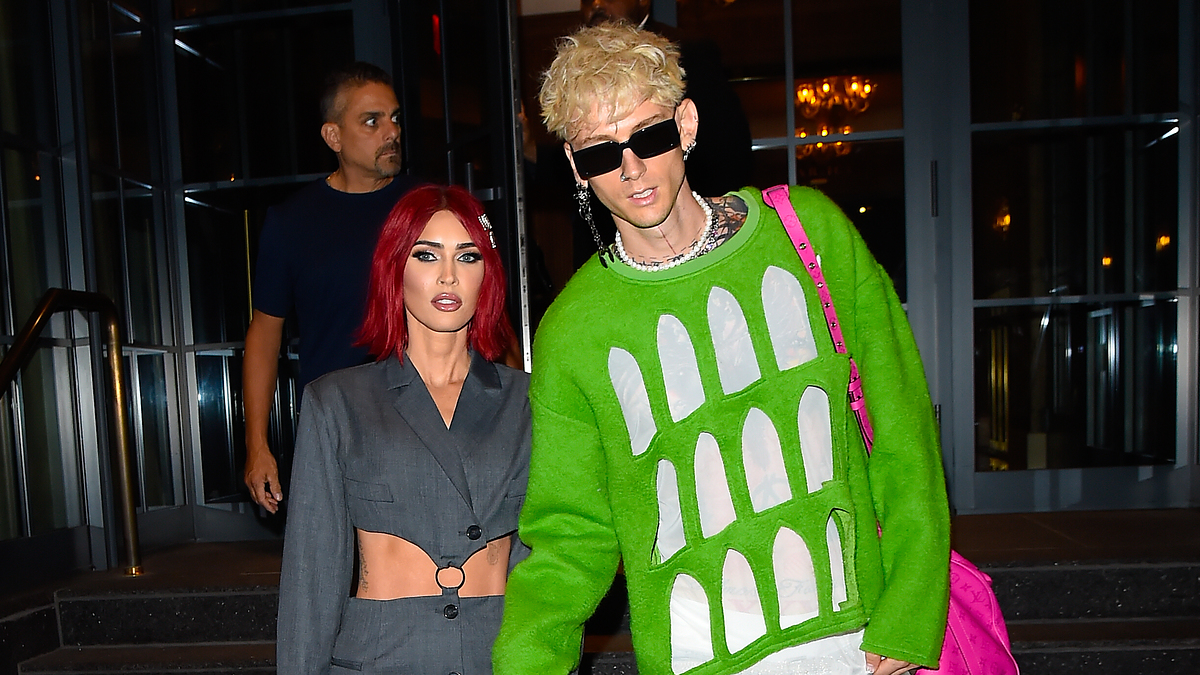 Megan Fox in a cropped blazer and skirt, and Machine Gun Kelly in a unique green sweater with cutouts and jeans, holding hands while walking outside a building