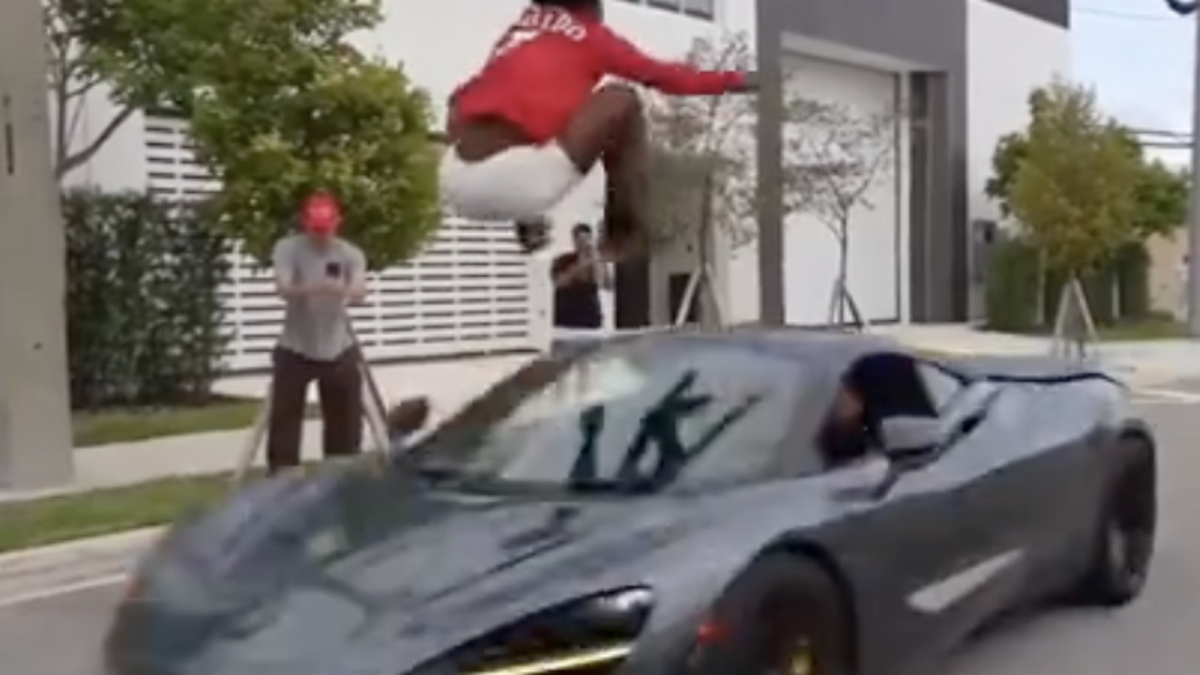Person in red hoodie jumps over a moving sports car on a city street while two others watch and film the scene