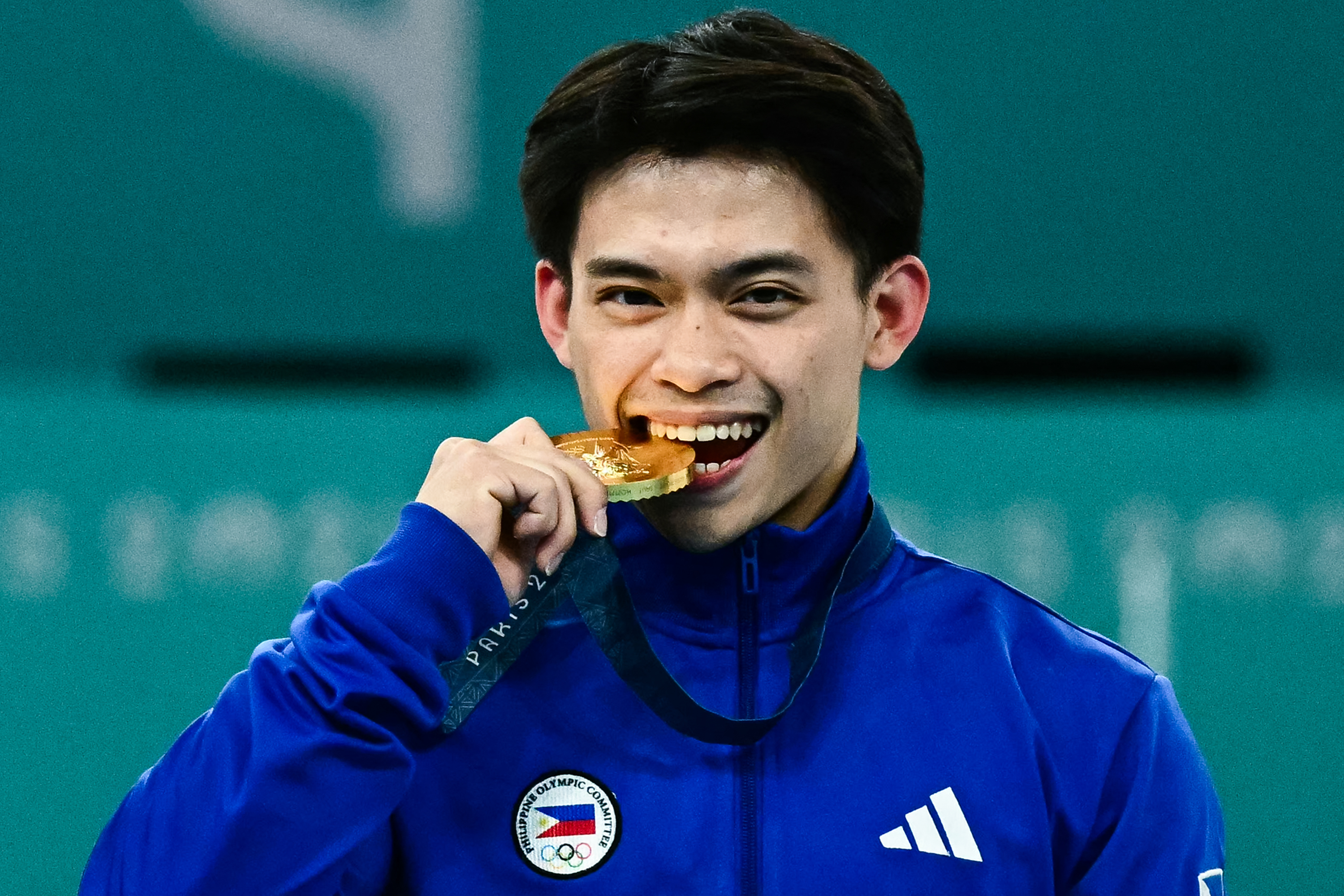 Carlos gnaws on an Olympic gold meda. He is in a blue jacket. 