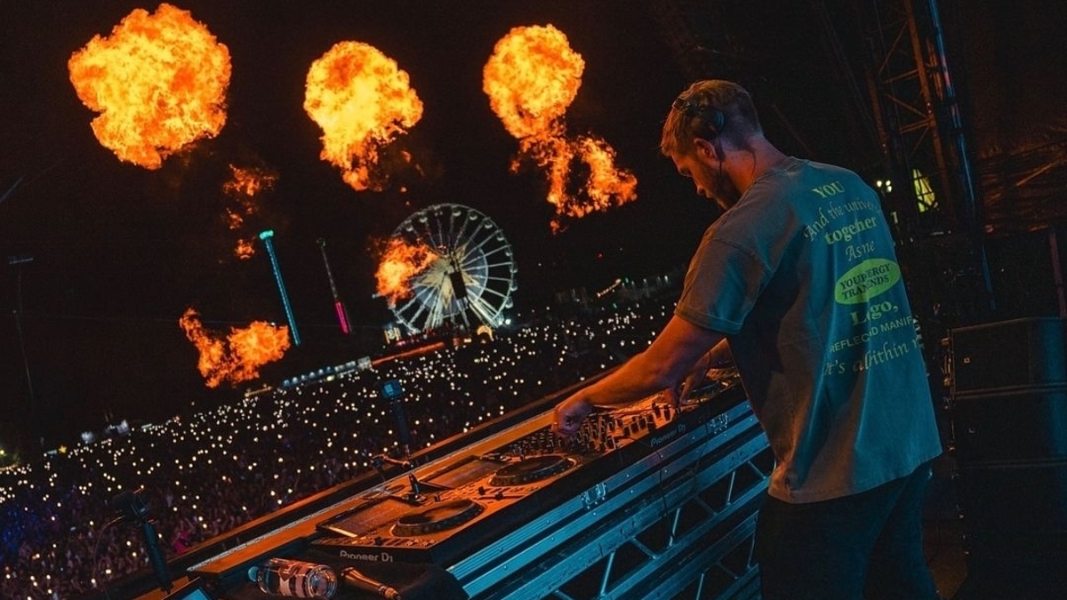 A DJ performs on stage at a music festival with flames erupting in the background and a large crowd in front of a Ferris wheel