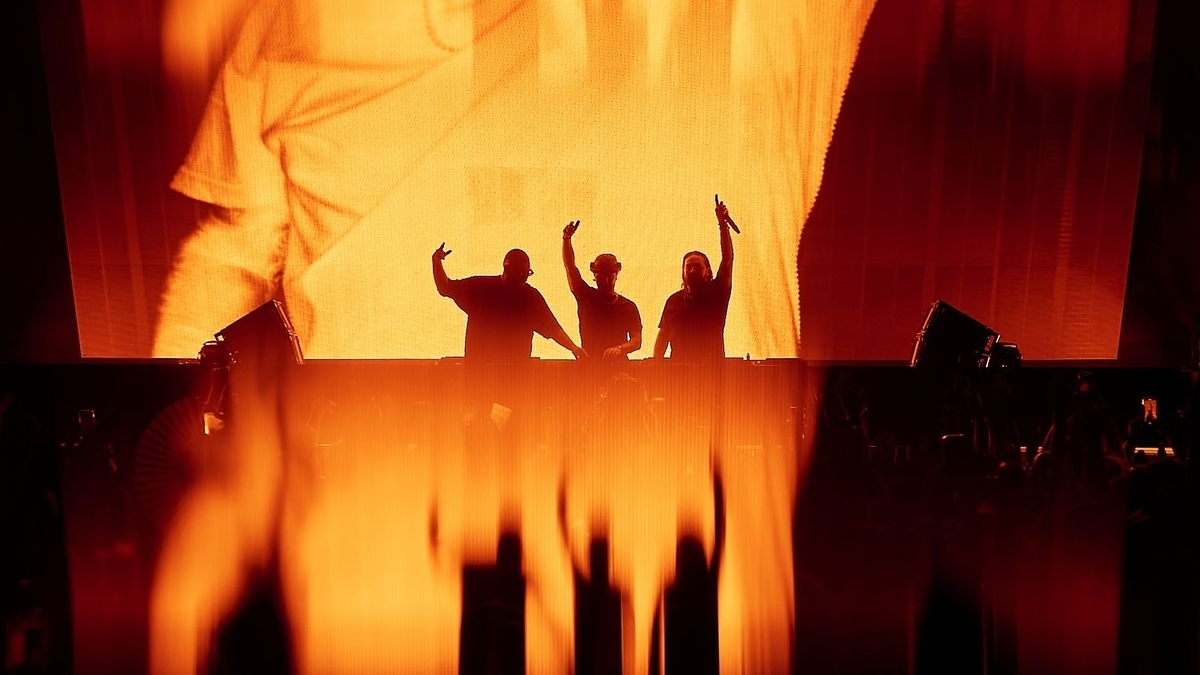 Three people, standing on stage with microphones, silhouettes against a large display screen with a face image in the background