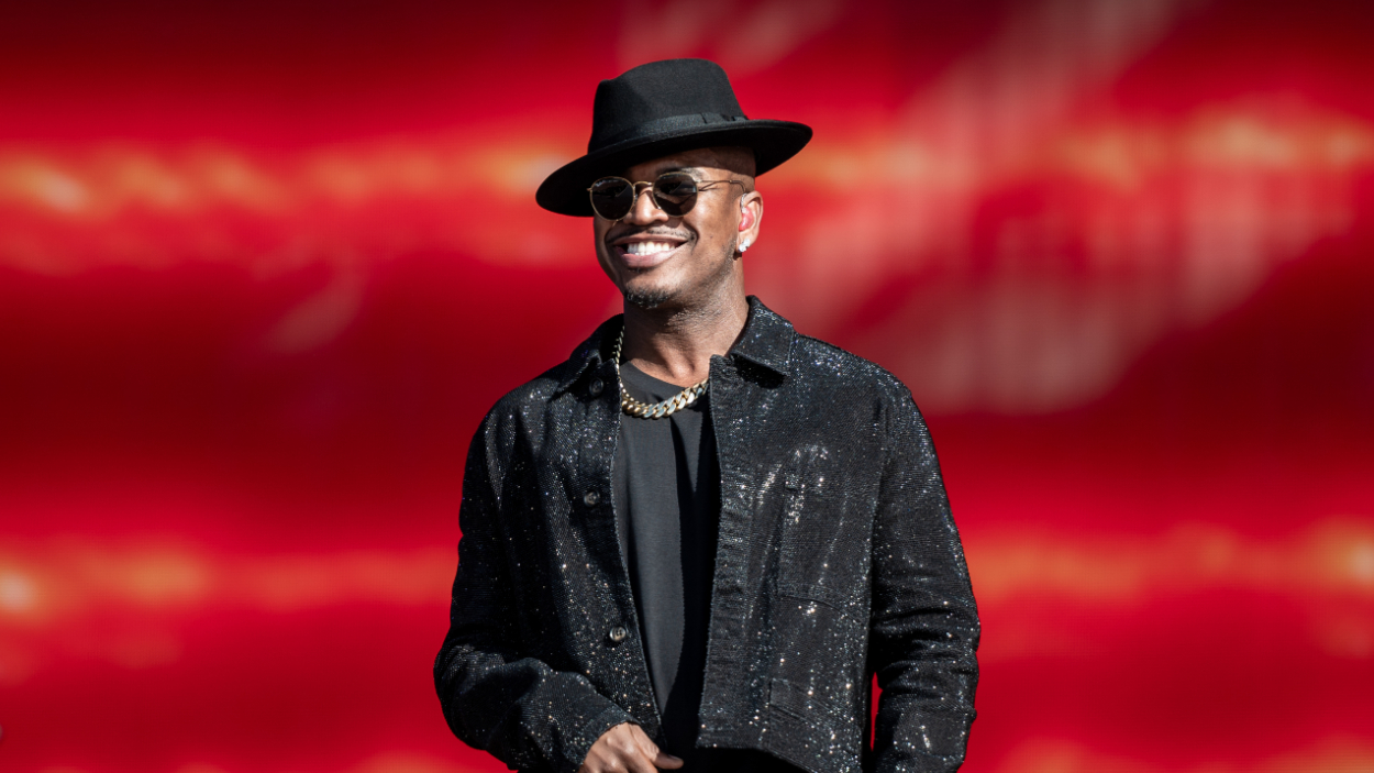 Ne-Yo performs on stage wearing a long-brimmed fedora hat, chain necklace, and black ensemble