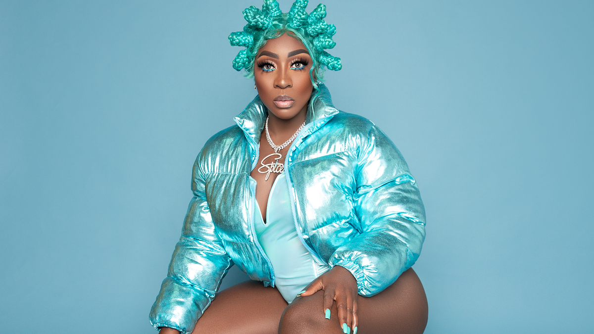 A woman wearing an iridescent puffer jacket, matching headpiece, leotard, and platform boots poses against a blue background