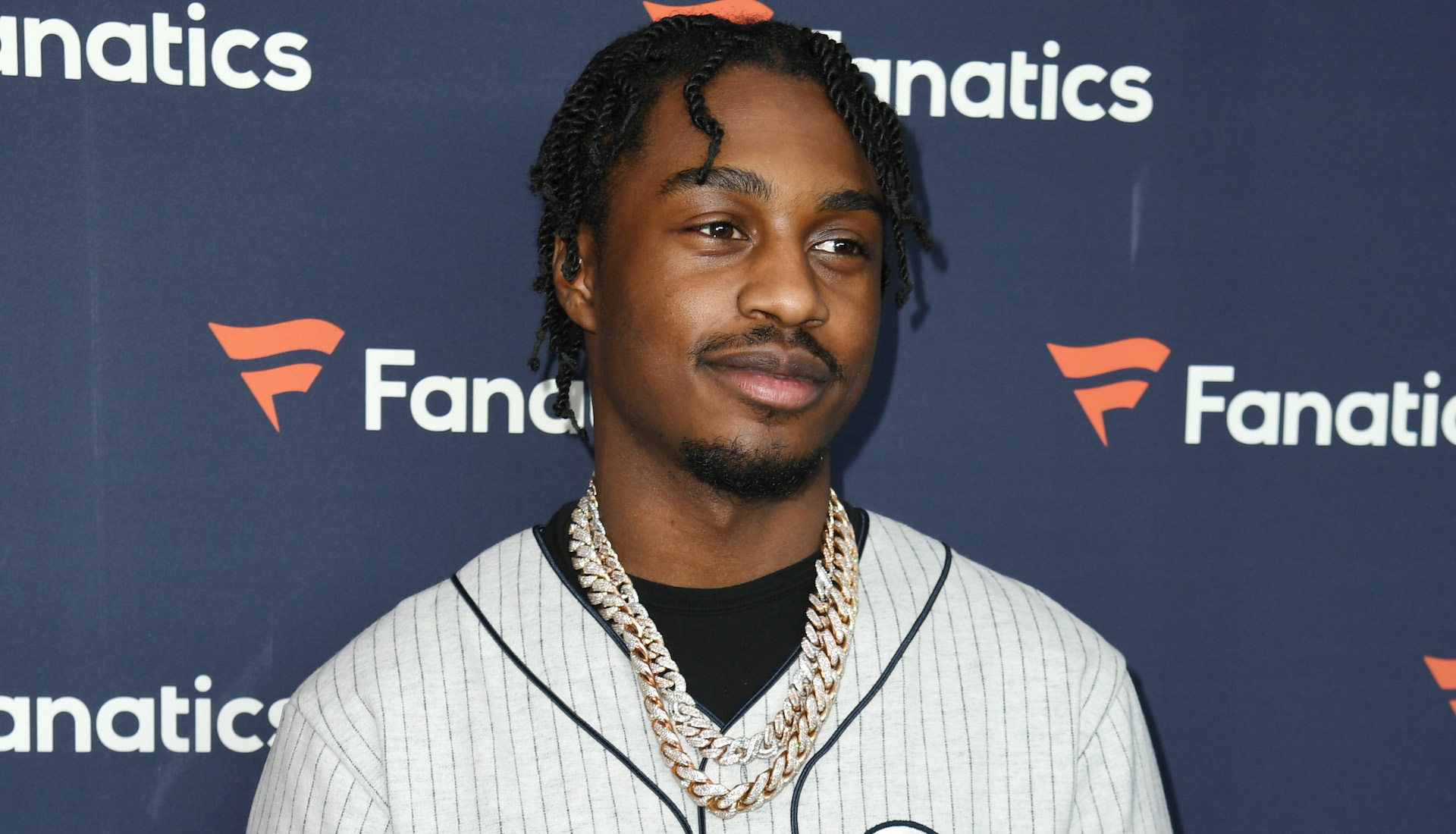 A person wearing a gray jersey and gold chains, standing in front of a backdrop with "Fanatics" logos.