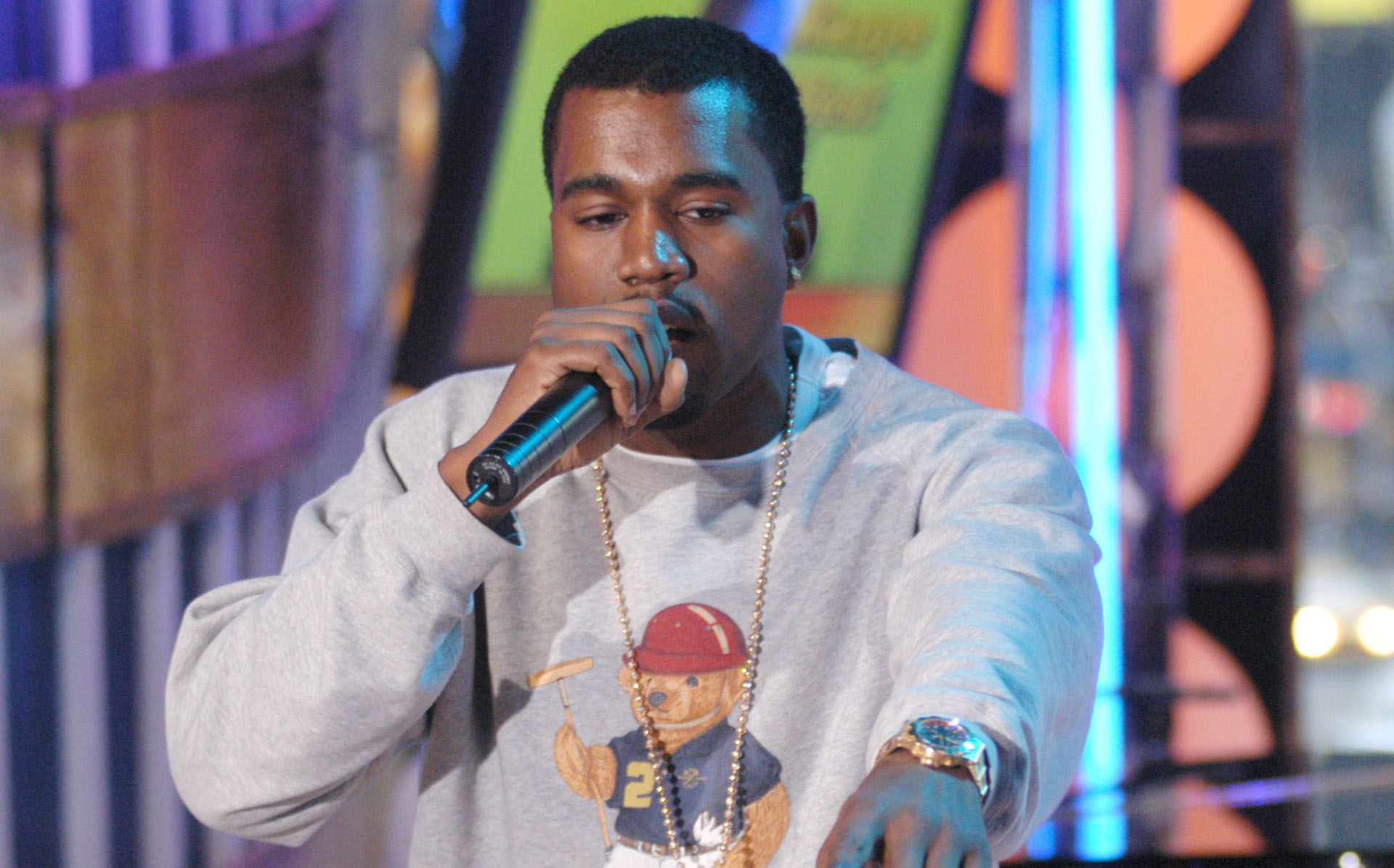 Kanye West performing on stage, wearing a gray sweatshirt with a bear graphic, holding a microphone.