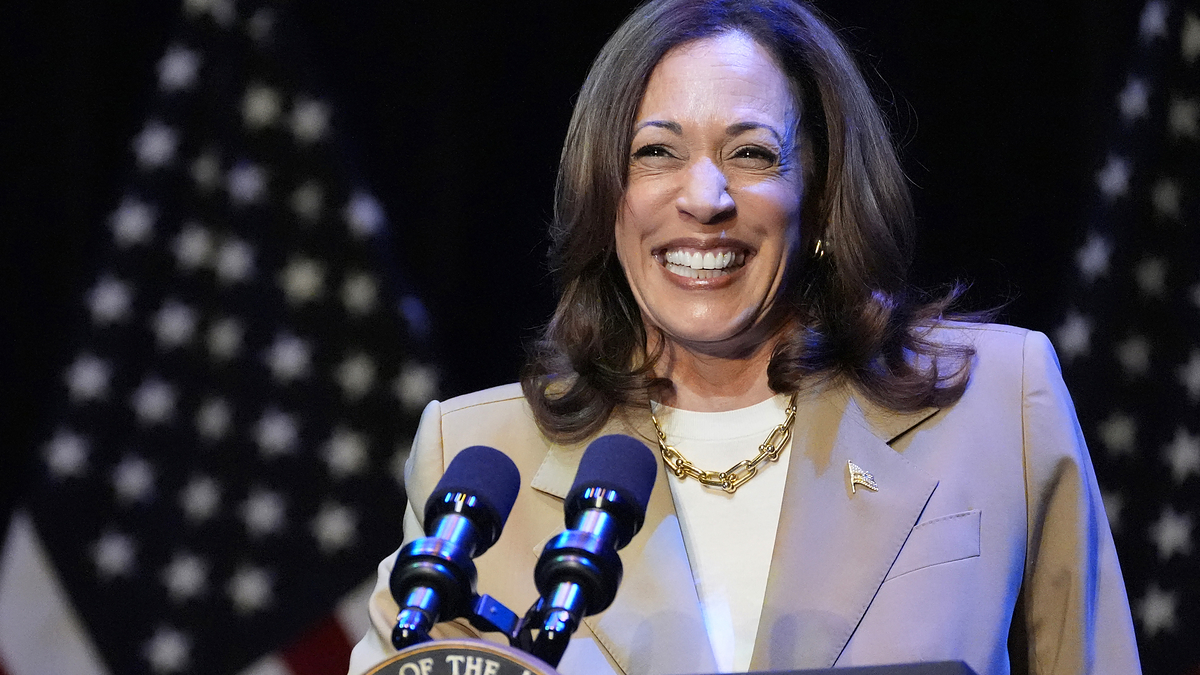 Kamala Harris smiles while speaking at a podium, two American flags in the background. She is wearing a beige blazer and a white shirt