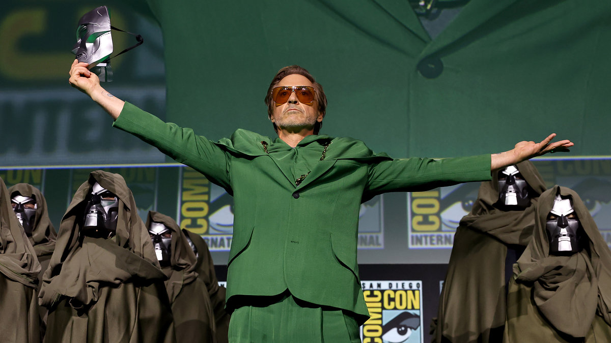 Robert Downey Jr. on stage at Comic-Con, wearing a green suit with arms outstretched, surrounded by people in hooded cloaks and metallic masks