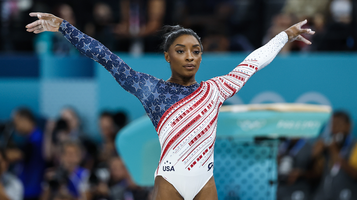 Simone Biles performs a gymnastics routine at the Olympics, wearing a leotard with stars and stripes. A podium is visible in the background