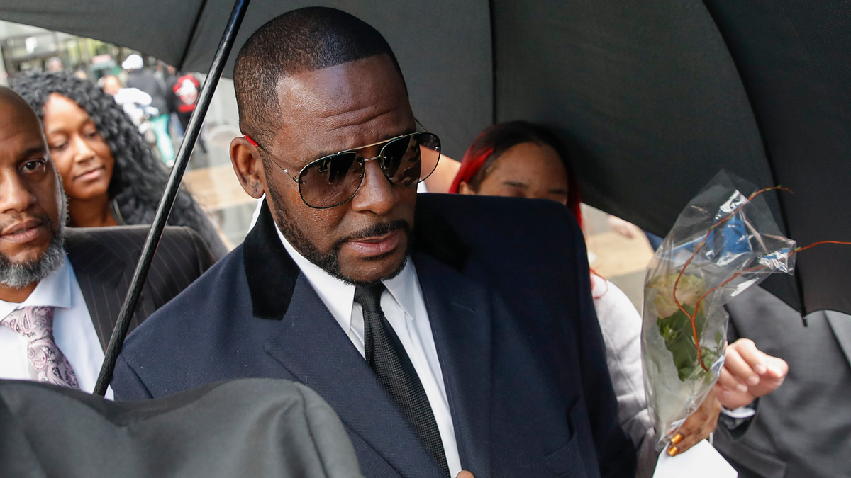 R. Kelly, holding an umbrella, is surrounded by people as he walks. He is wearing aviator sunglasses, a black suit, white shirt, and black tie