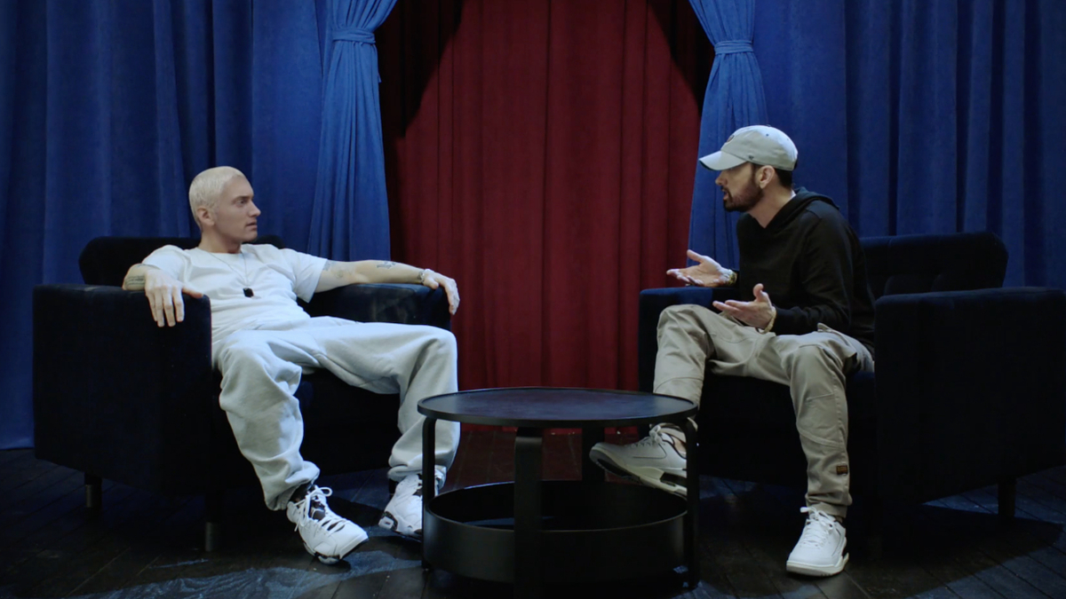 Eminem and Eminem impersonator sitting on chairs, engaged in conversation, with blue and red curtains in the background