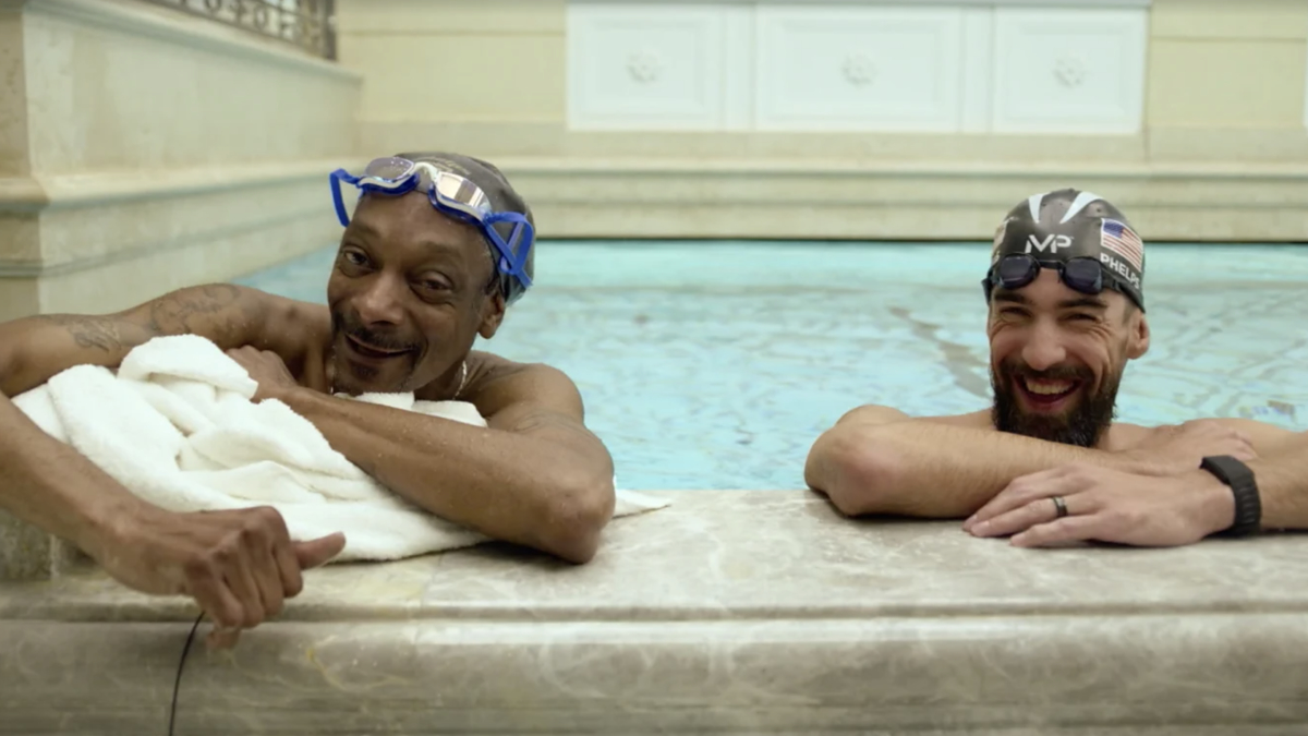 Snoop Dogg and Michael Phelps having a conversation while resting in a pool. Snoop Dogg wears blue swim goggles and holds a towel; Michael Phelps has a swim cap on