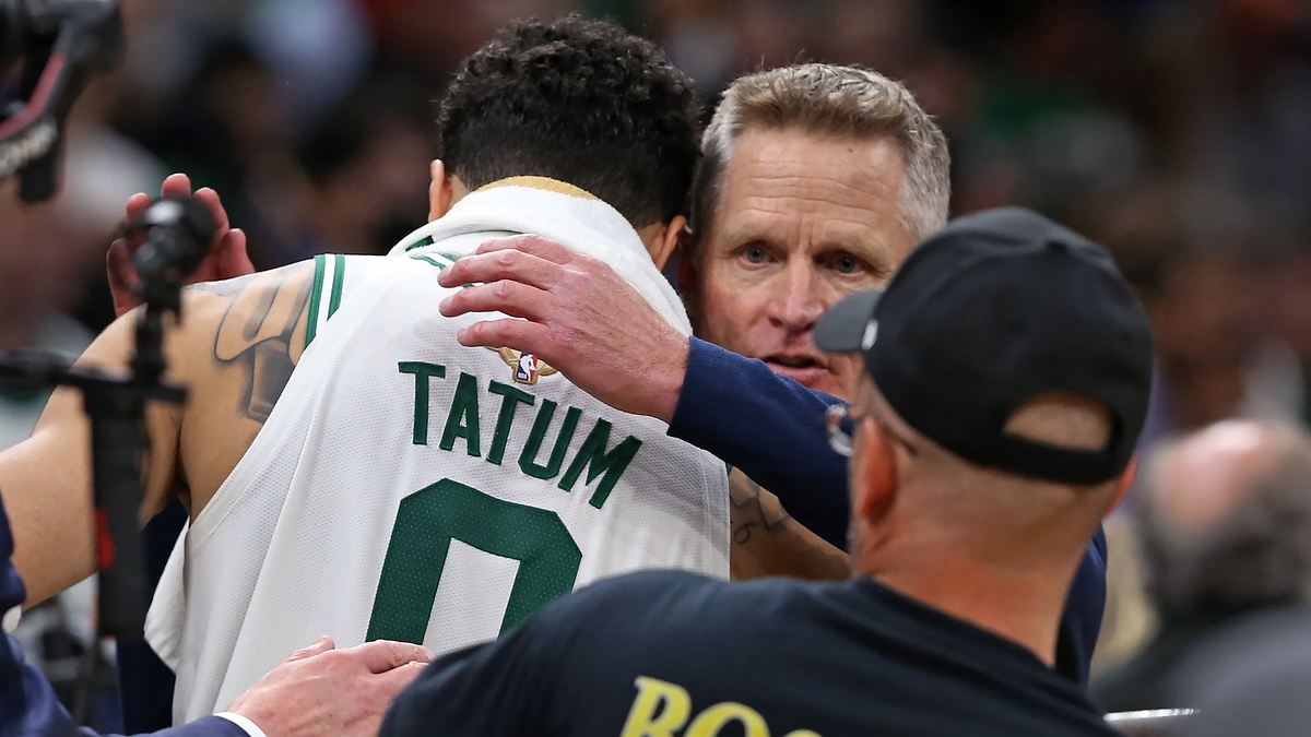 Jason Tatum of the Boston Celtics is embraced by a coach or staff member on the court during a basketball game. A man in a "Boston" shirt is also visible