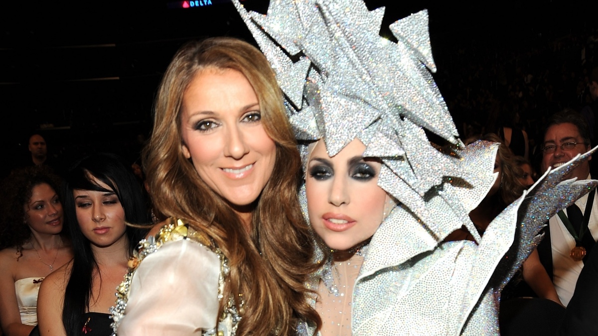 Celine Dion and Lady Gaga pose together. Dion wears a dress with gold details, while Gaga wears an avant-garde outfit with an elaborate headdress