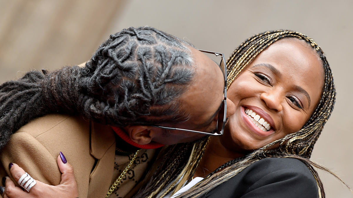 Two people, one with long dreadlocks in a tan jacket and the other smiling woman with long braids in a black blazer, sharing a close, joyful moment