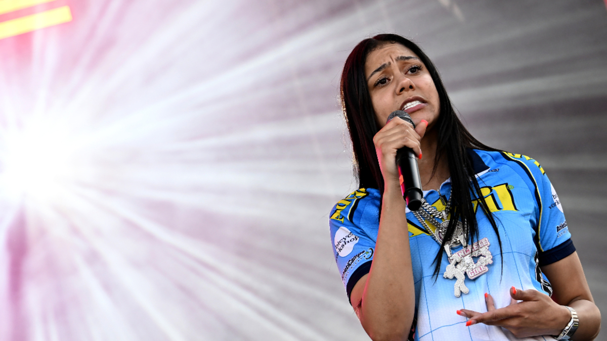 A woman on stage holds a microphone and sings passionately, wearing a blue-themed sports jersey with an elaborate necklace, against a radiant light background