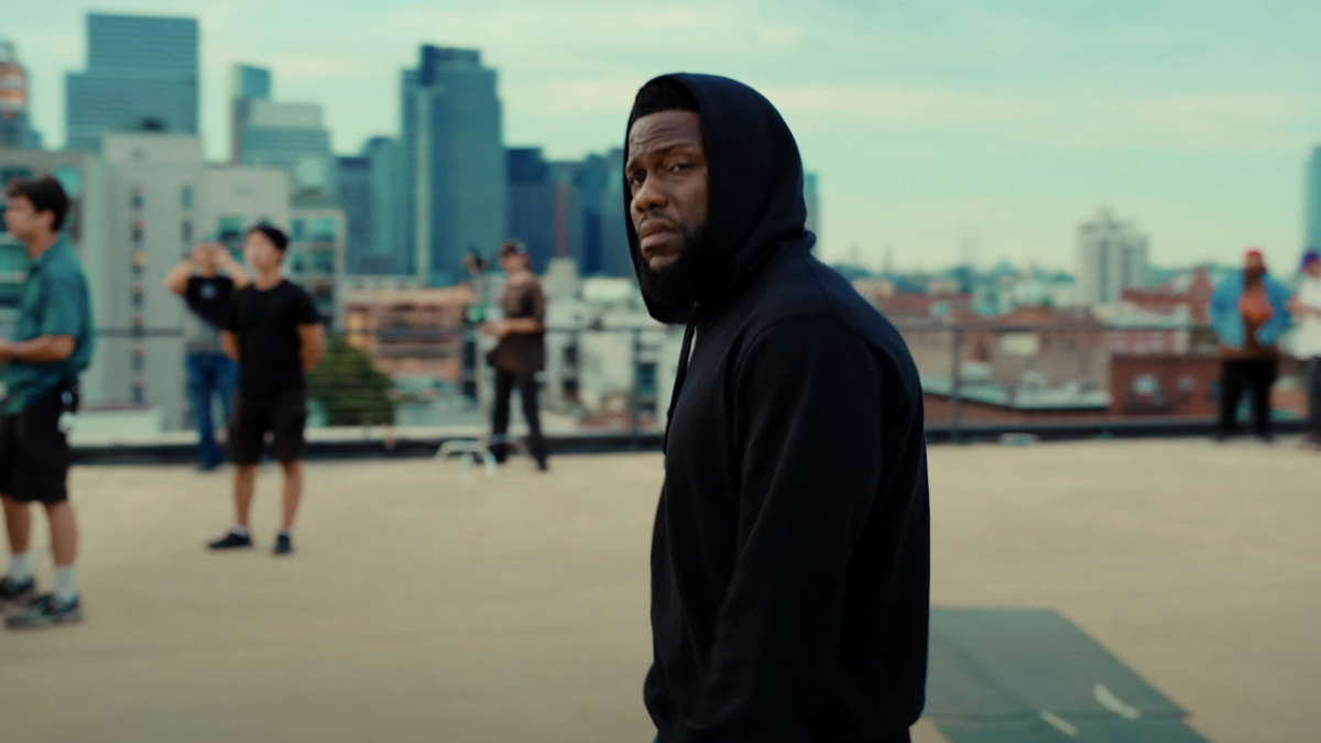 Kevin Hart stands on a rooftop in a cityscape, wearing a black hoodie. Crew members and photographers are in the background