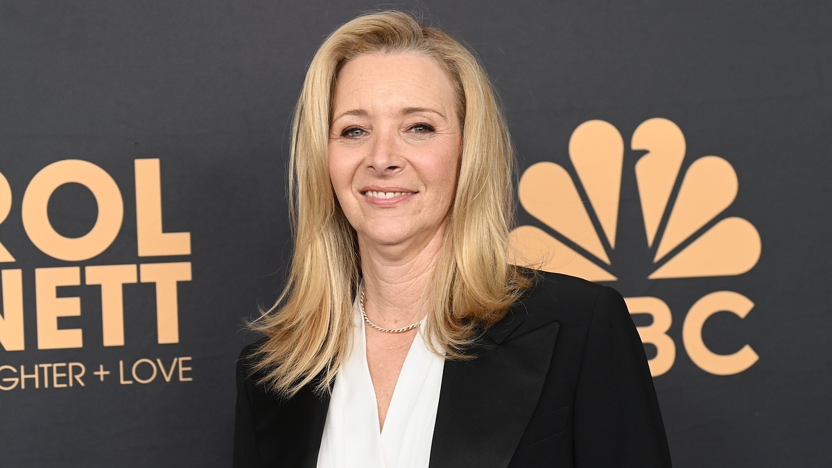 Lisa Kudrow in a black jacket and white shirt at the Carol Burnett: 90 Years of Laughter + Love event, posing in front of an NBC logo backdrop