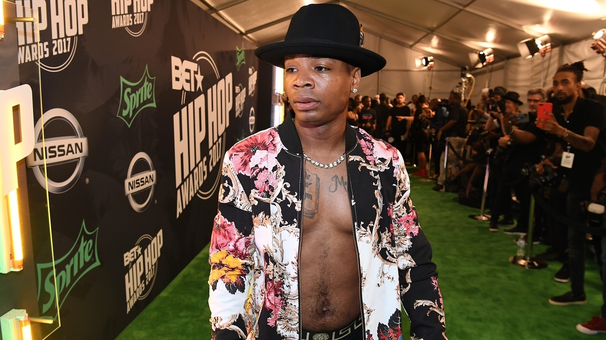 Plies on the BET Hip Hop Awards 2017 red carpet, wearing an open floral jacket, black hat and multiple necklaces, surrounded by photographers