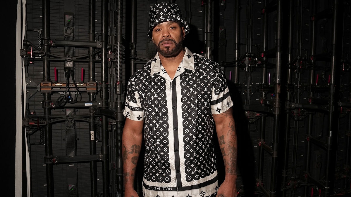 Man in a Louis Vuitton patterned shirt and matching hat stands in front of a high-tech equipment backdrop