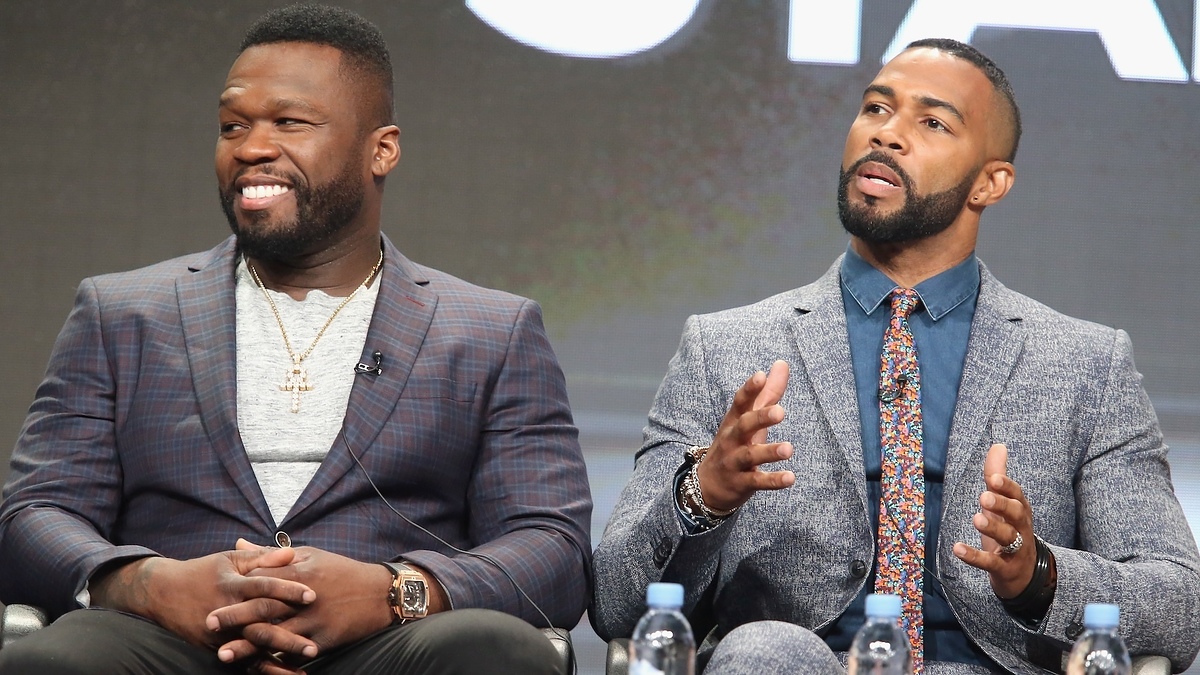 50 Cent wearing a checked blazer and gray shirt, and Omari Hardwick in a patterned tie and textured suit, talking while seated at a panel discussion