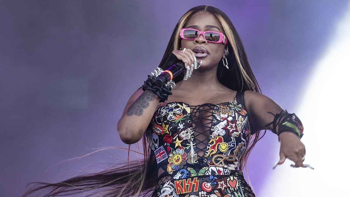 A woman in a patchwork bustier top, pink glasses, and long hair performs on stage, holding a microphone