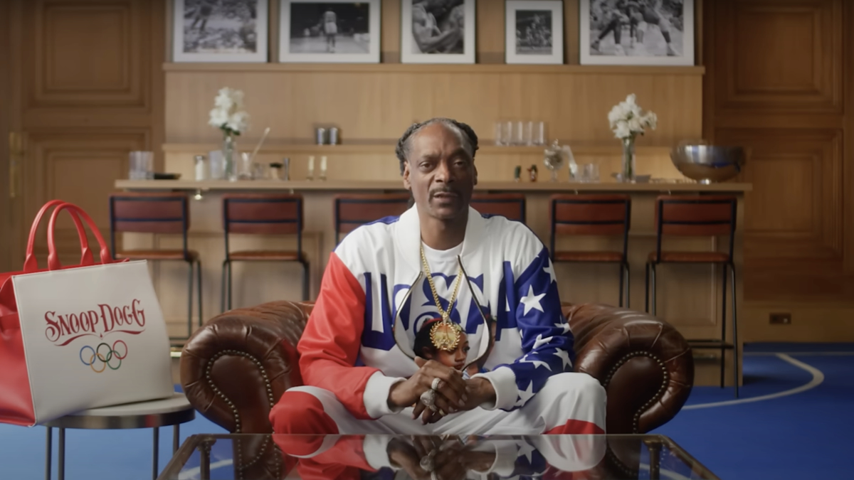 Snoop Dogg sitting on a leather couch in a stylish room, wearing a patriotic tracksuit and multiple gold chains. A "Snoop Dogg Olympic" bag is nearby