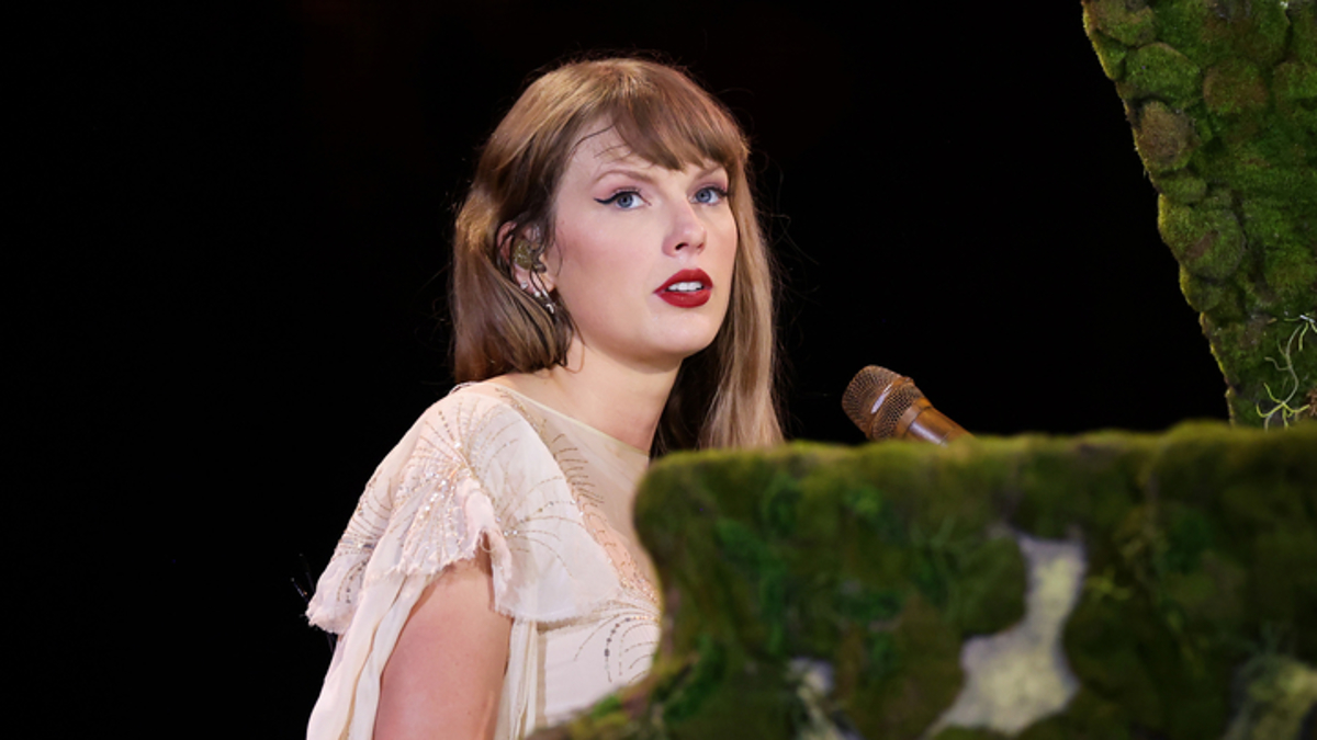 Taylor Swift performs at a moss-covered piano, wearing a flowing cream-colored dress. She is focused, singing into a gold microphone