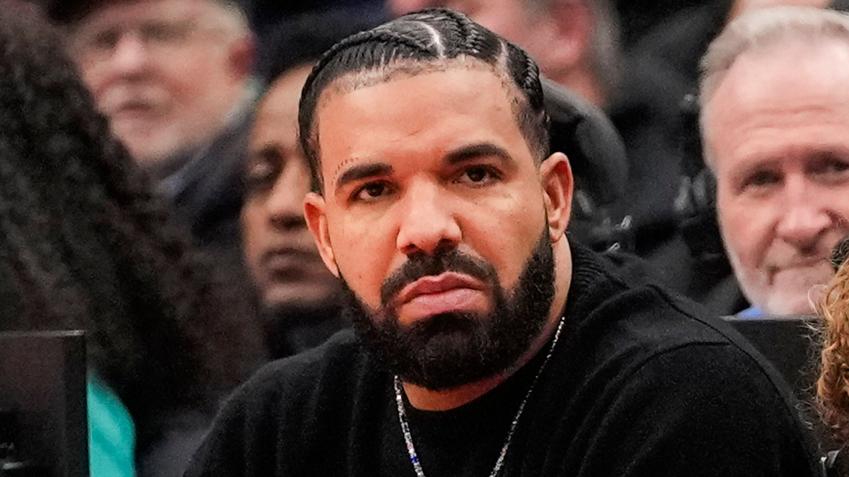 Drake sits in the audience at an event, wearing a black sweater and a necklace, with braided hair and a trimmed beard