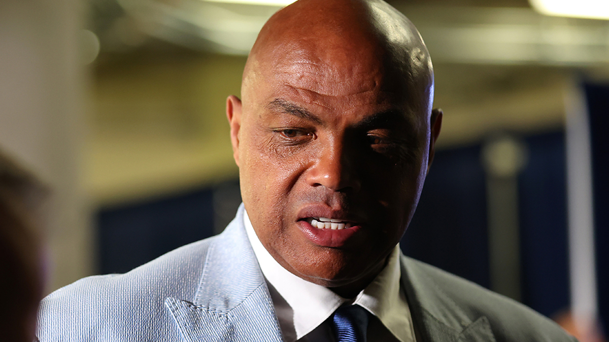 Charles Barkley in a light suit and tie, speaking with someone at a sports event