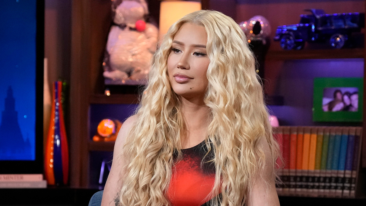 Iggy Azalea with long curly hair, wears casual attire in a studio set with colorful decorations and a bookshelf in the background