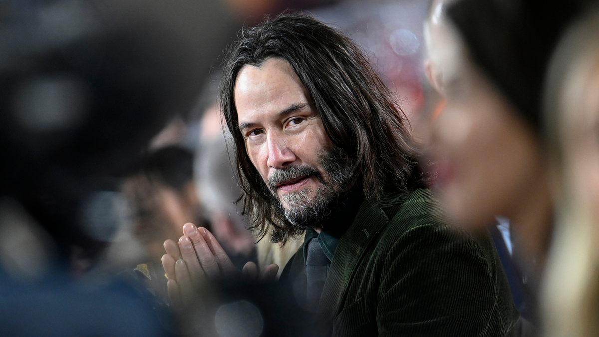 Keanu Reeves with shoulder-length hair and beard, wearing a dark suit, at a public event, hands clasped