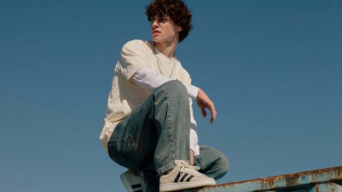 Young person with curly hair wearing casual clothing and Adidas sneakers sits on top of a shipping container, looking off into the distance