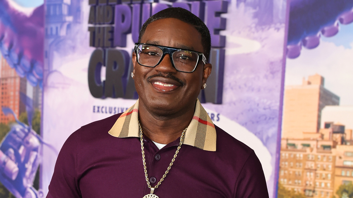 Person smiling at the camera on a movie premiere background. The person is wearing glasses, a maroon shirt with a distinctive plaid collar, and a large chain necklace