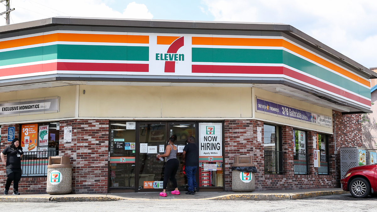 People stand outside a 7-Eleven convenience store with a "Now Hiring" sign visible on the right
