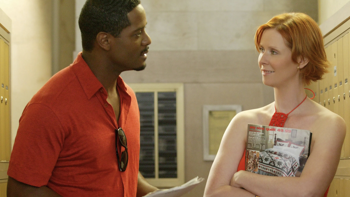 Blair Underwood, in a casual shirt, talks to Cynthia Nixon, who is holding magazines and wearing an elegant sleeveless dress, in a hallway with lockers