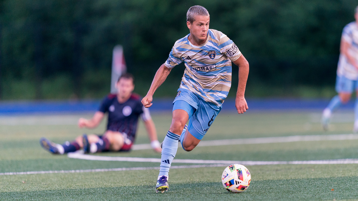 A soccer player in mid-action, wearing a striped jersey and blue shorts, dribbles the ball on the field as a defender sits on the ground in the background