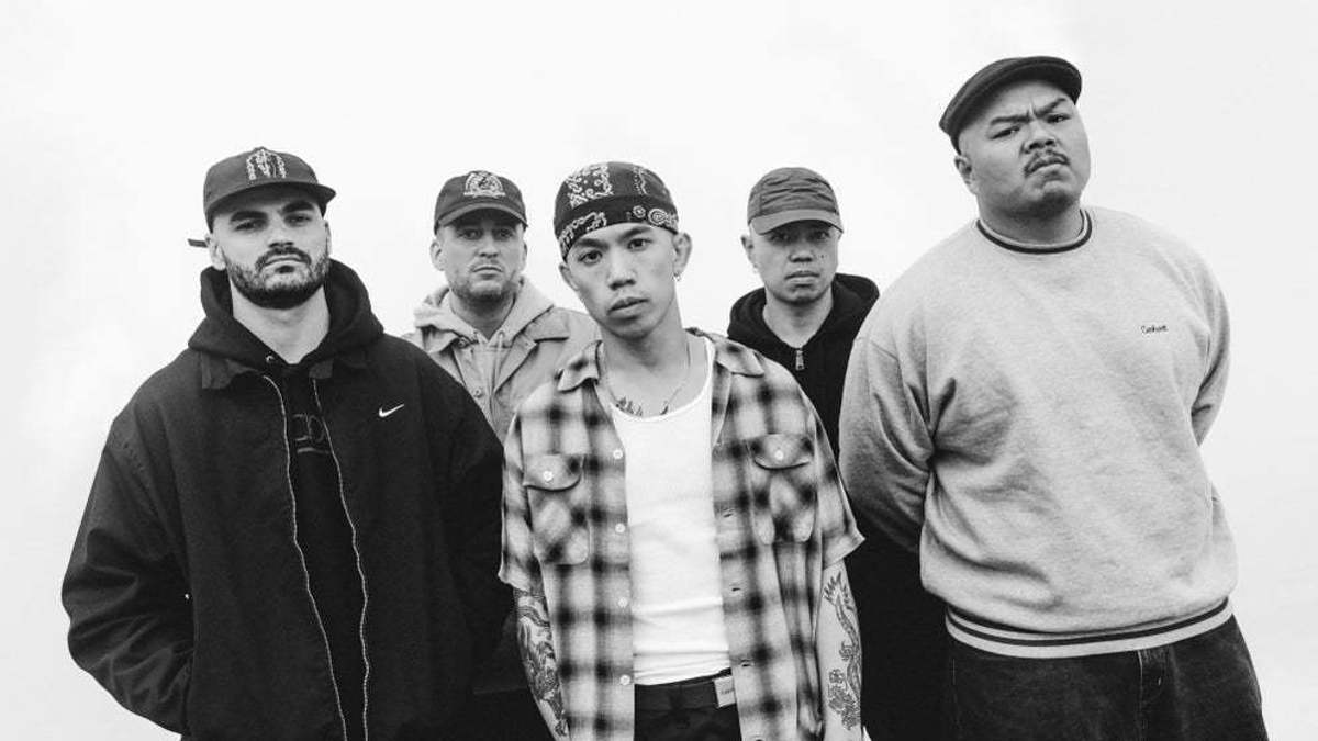 The image shows five members of a musical group posing together. They are dressed in casual streetwear, featuring hoodies, caps, and a plaid shirt