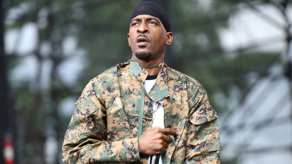 Rakim in a camouflage jacket and black headwrap performs on stage, microphone in hand, with an expressive look on his face