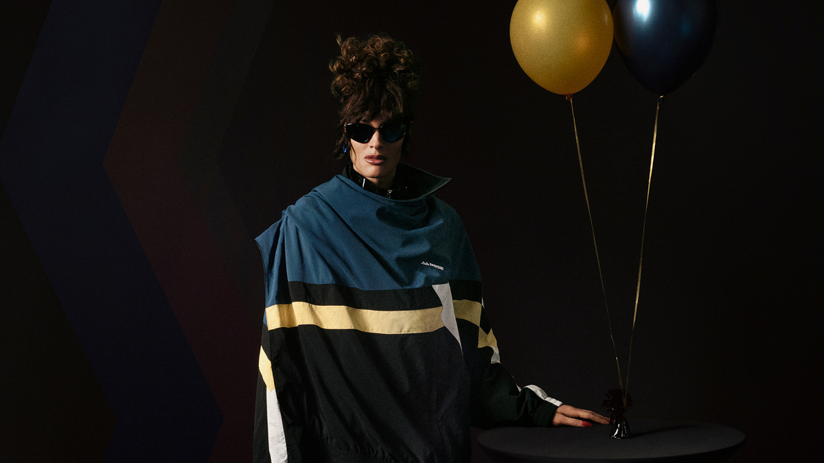 Person in oversized, sporty jacket with a high collar and black shades standing next to a table with gold and black balloons against a dark backdrop. Names unknown